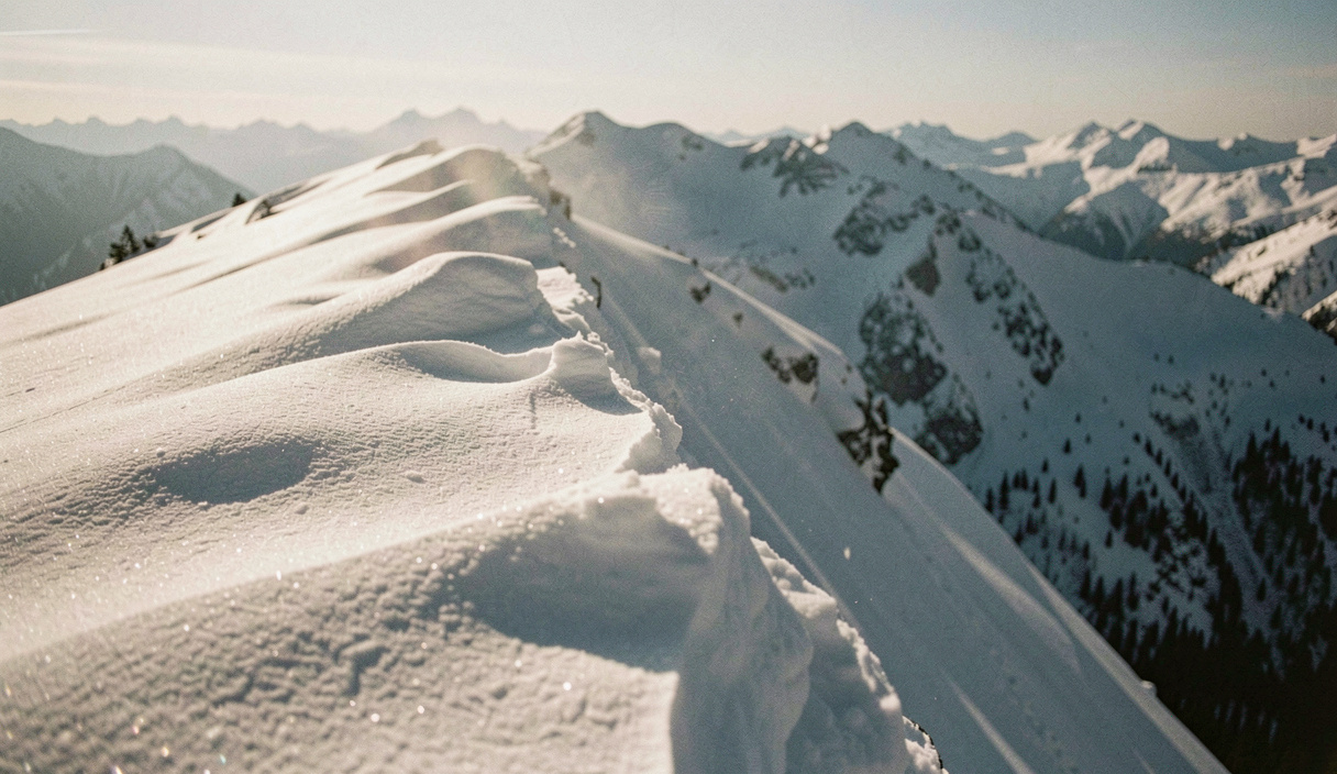 Vom Wind geformte Wechte auf einem Grat mit frischen Triebschneepolstern darunter, Schneefahnen wehen über die Kante, hartes Wintermorgenlicht, sehr breite Aufnahme aus mittlerer Distanz